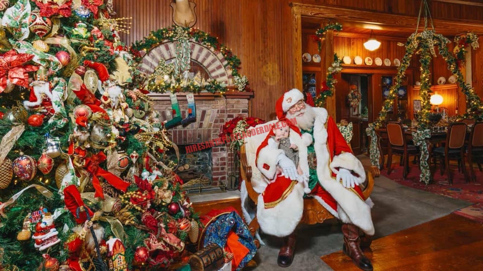 Professional Santa for hire in Broward seated by a fireplace and tree holding a baby