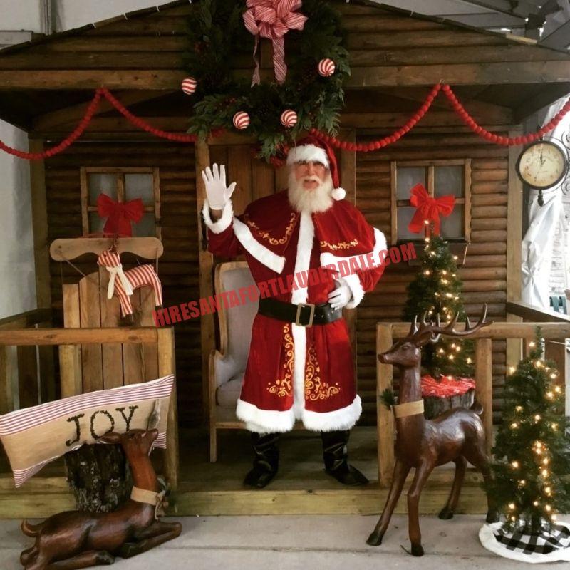 Real bearded Santa for hire waving from a log-cabin doorway with wooden reindeer